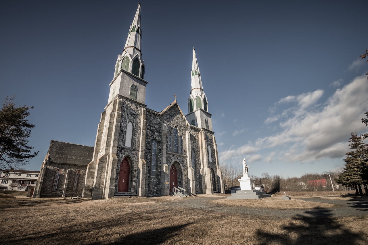 Masonry Catholic church against trees and statue on pedestal under cloudy blue sky in sunlight